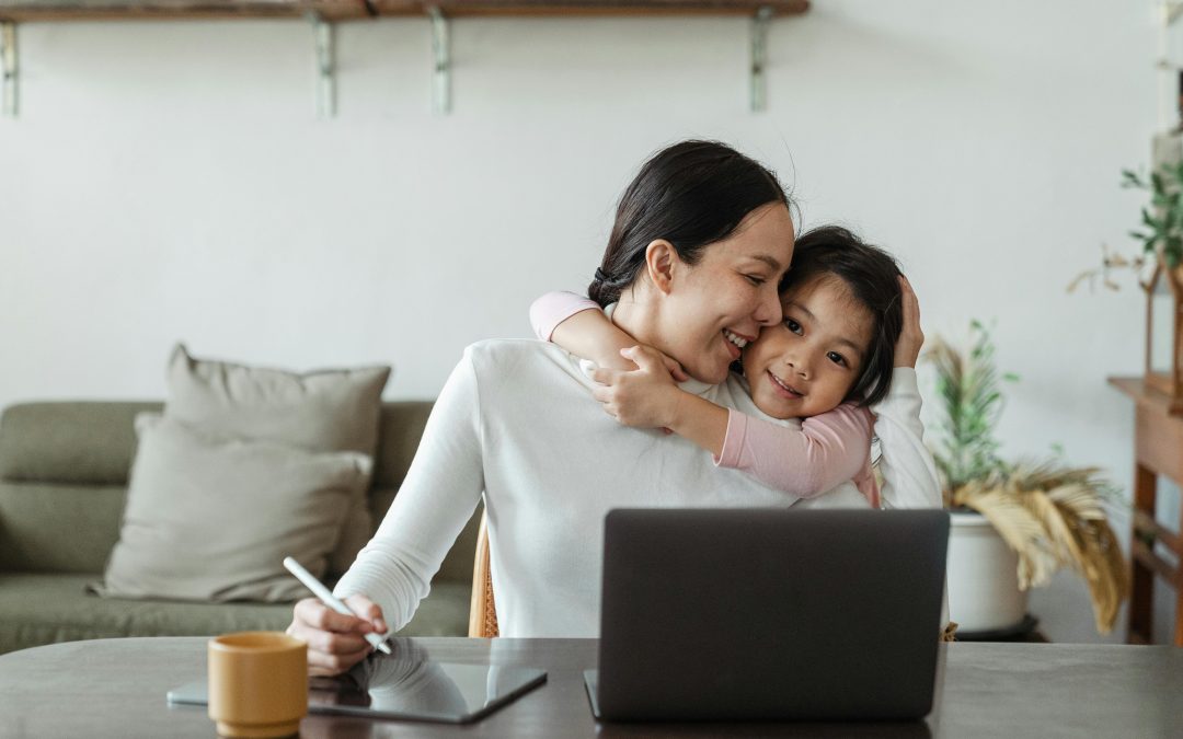 woman who feels confident in her skin and is working at a table on a laptop while a child hugs her from behind