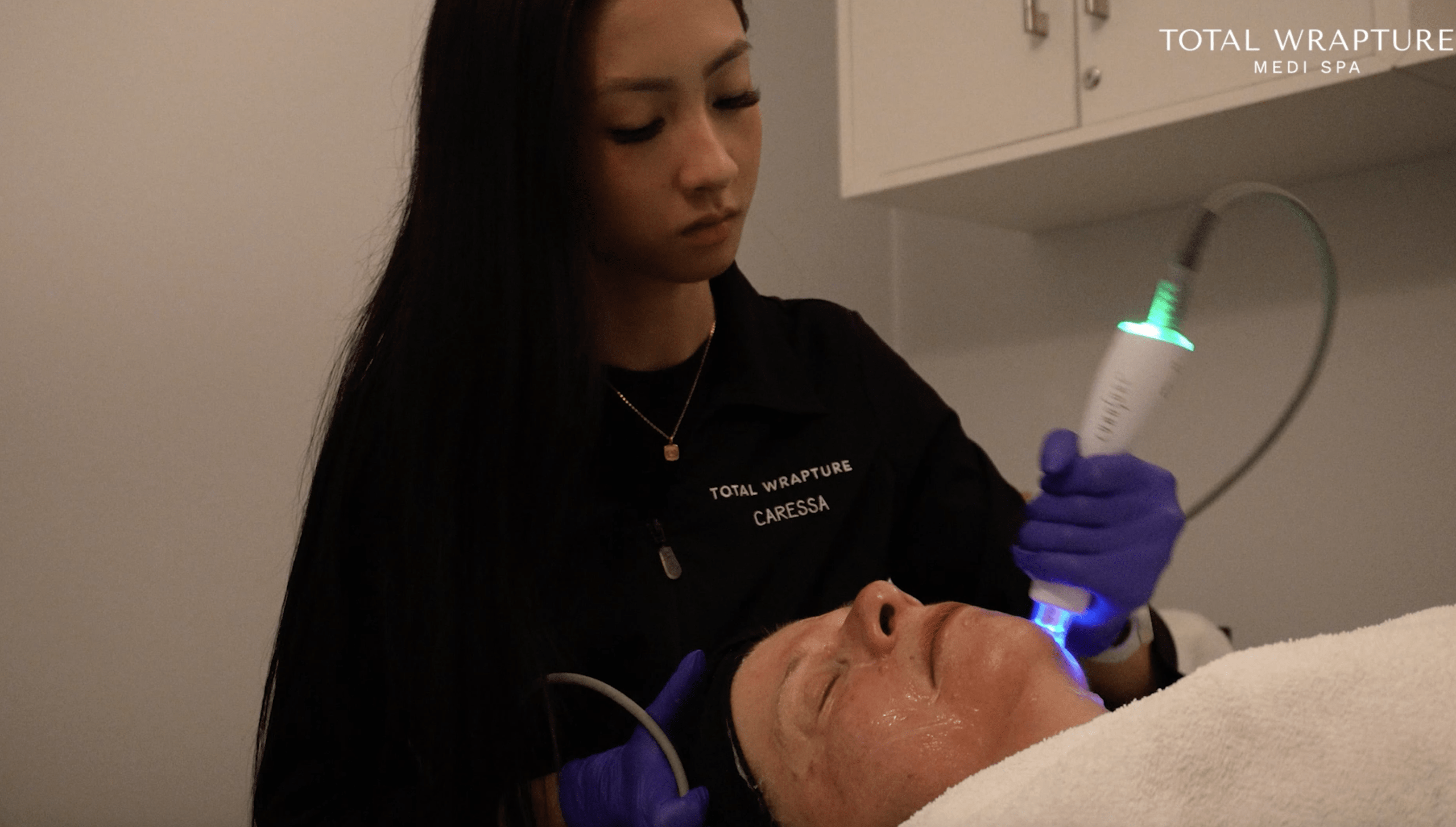 Esthetician in a black smock giving a facial RF skin tightening treatment to a woman lying down.