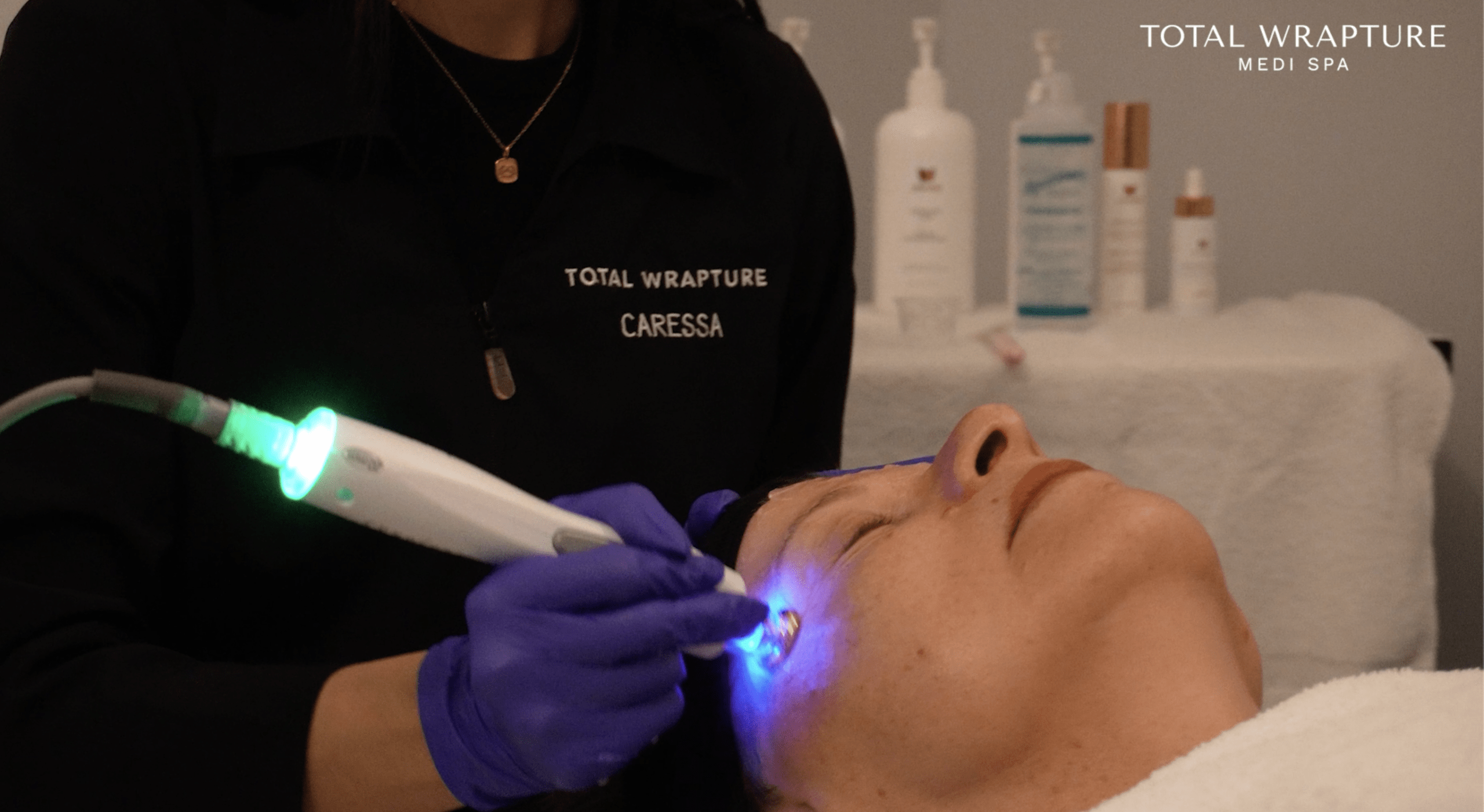 Esthetician in a black smock giving a facial RF skin tightening treatment to a woman lying down.